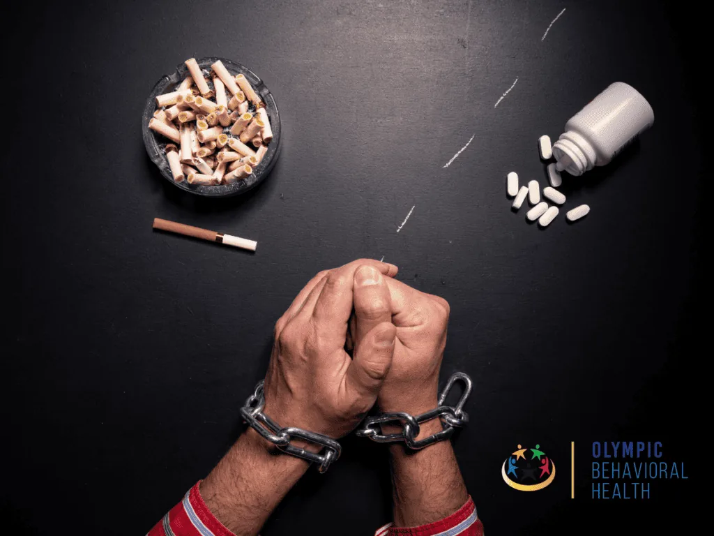 Hands bound by a chain beside an ashtray of cigarette butts and spilled pills, showing addiction's hold on the body