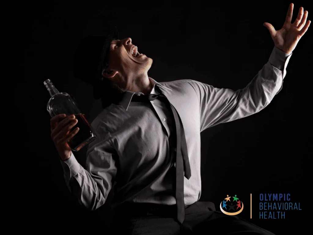 Man in a dark room holding a liquor bottle and shouting with arm raised, illustrating the symptoms of addiction