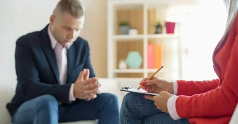 Man in a blazer clasping his hands while a therapist takes notes, illustrating anger management therapy in Lantana, FL