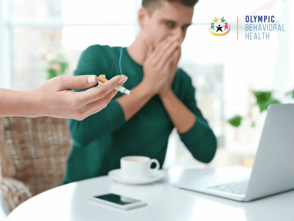 Man at a laptop covering his mouth while a hand offers him a cigarette, illustrating causes of nicotine addiction