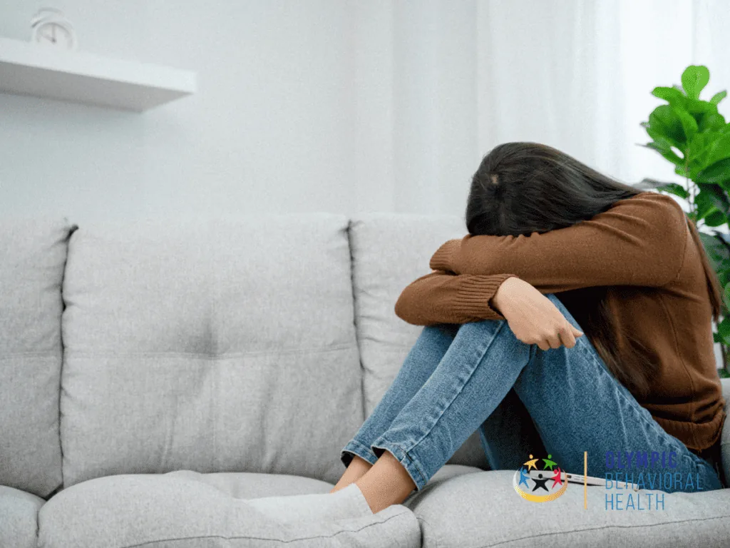 Young woman sitting on a gray sofa hugging her knees with her head down, illustrating a co-occurring disorder