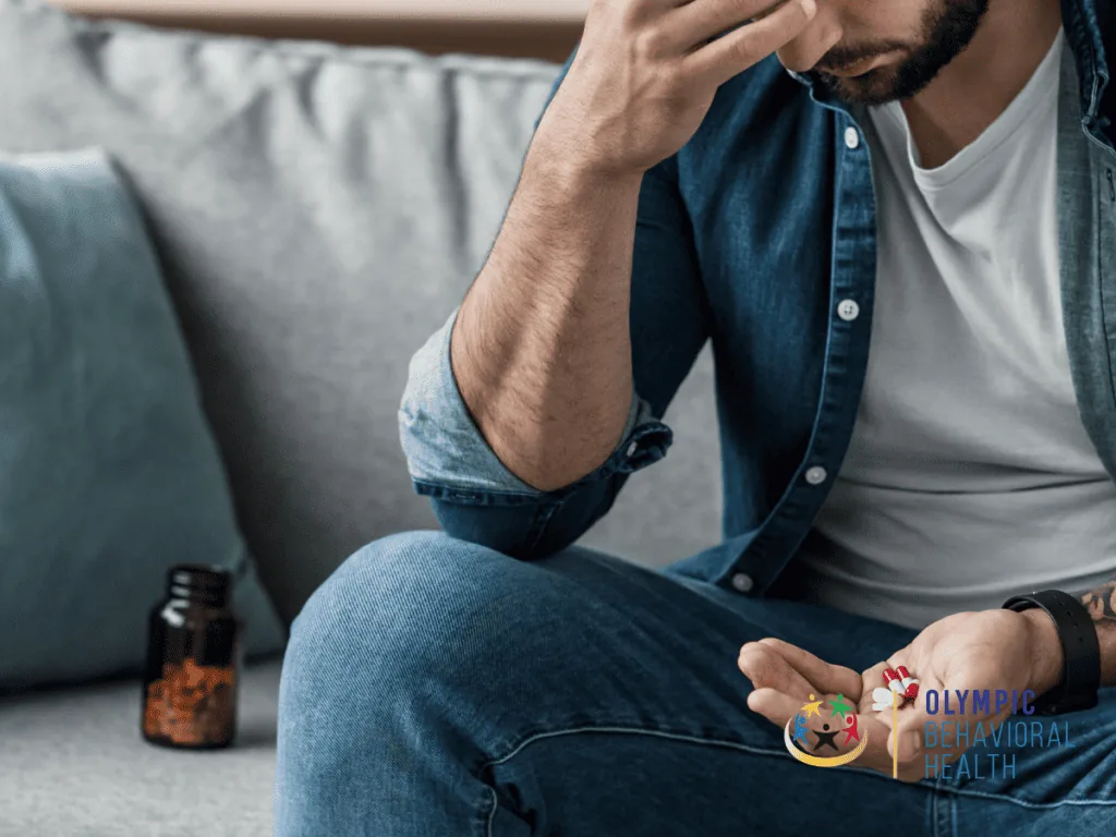 Man sitting on a couch with head in hand holding pills from a bottle, illustrating drug addiction and codependency