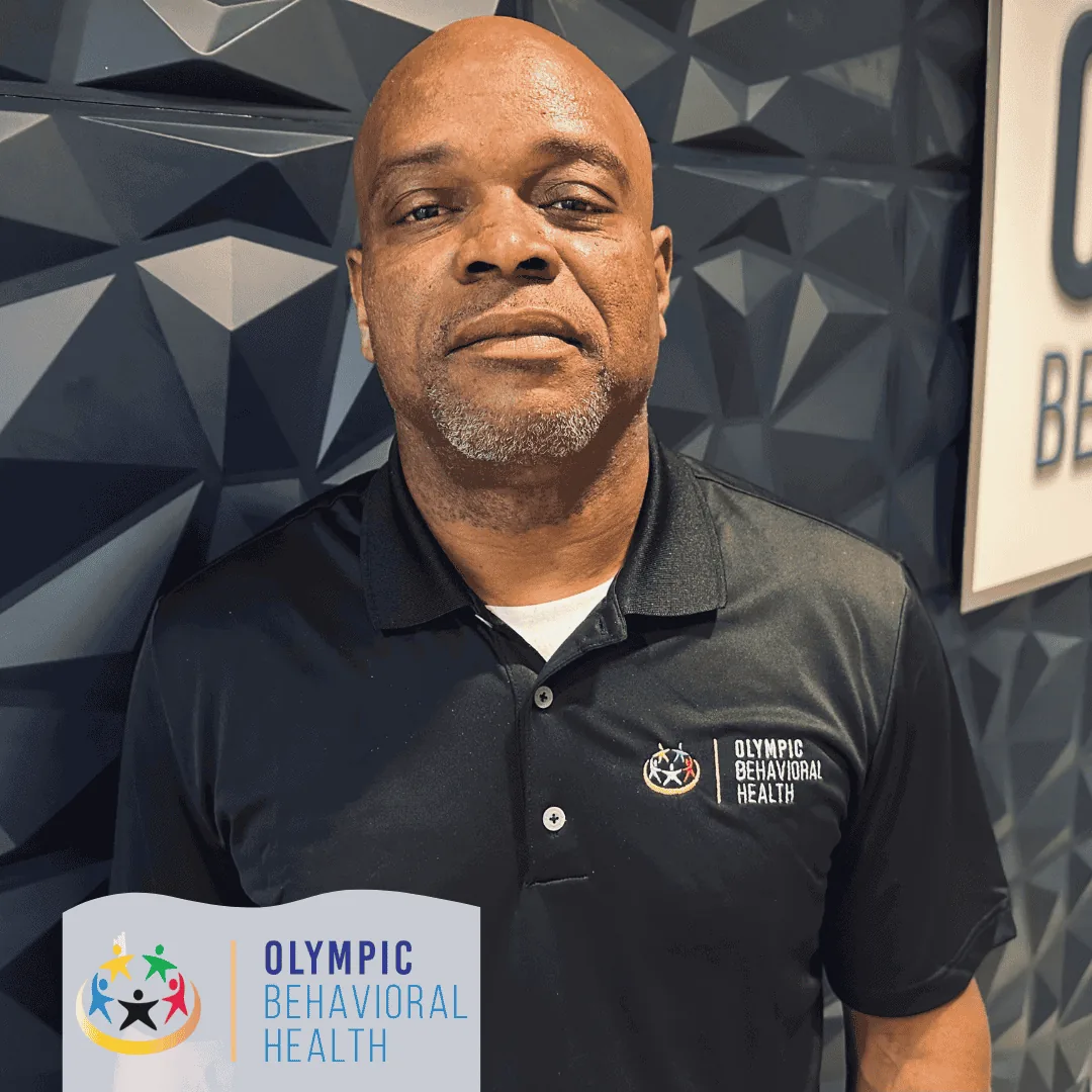 Jonathan Pollard, Alumni Coordinator at Olympic Behavioral Health, in a black branded polo in front of the logo wall