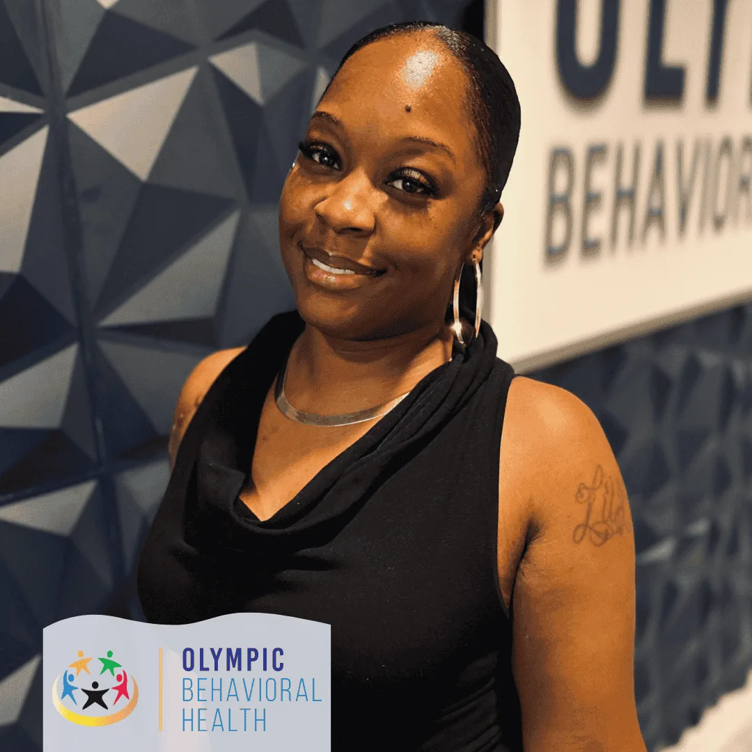 LaChondria Jackson, Lead Behavioral Health Technician at Olympic Behavioral Health, smiling by the logo wall