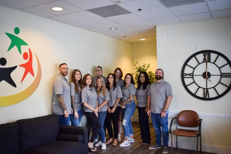 Olympic Behavioral Health staff in matching gray shirts posing in the lobby beside the logo mural and a large wall clock