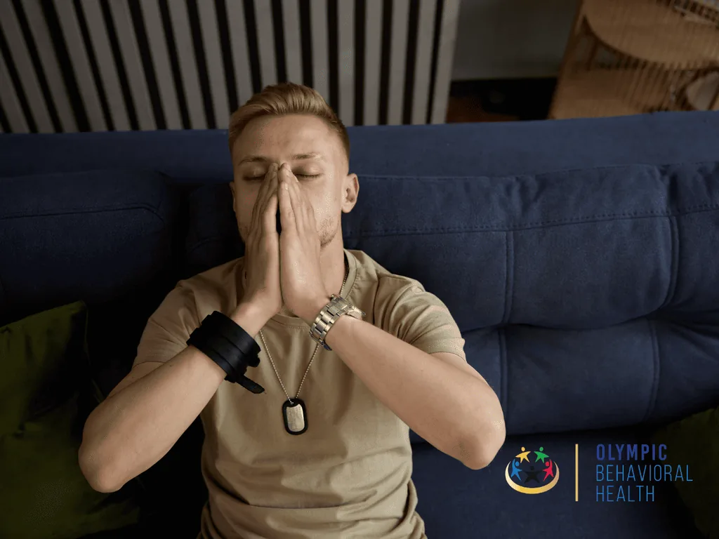Young man with dog tags sitting on a couch, eyes closed and hands pressed to his face, illustrating PTSD risk in veterans